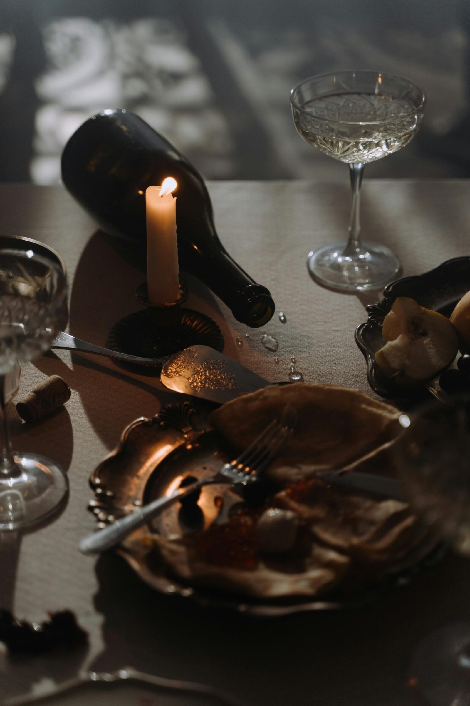 Moody candlelit table with wine bottle, empty plates, and scattered tableware, evoking a finished meal.